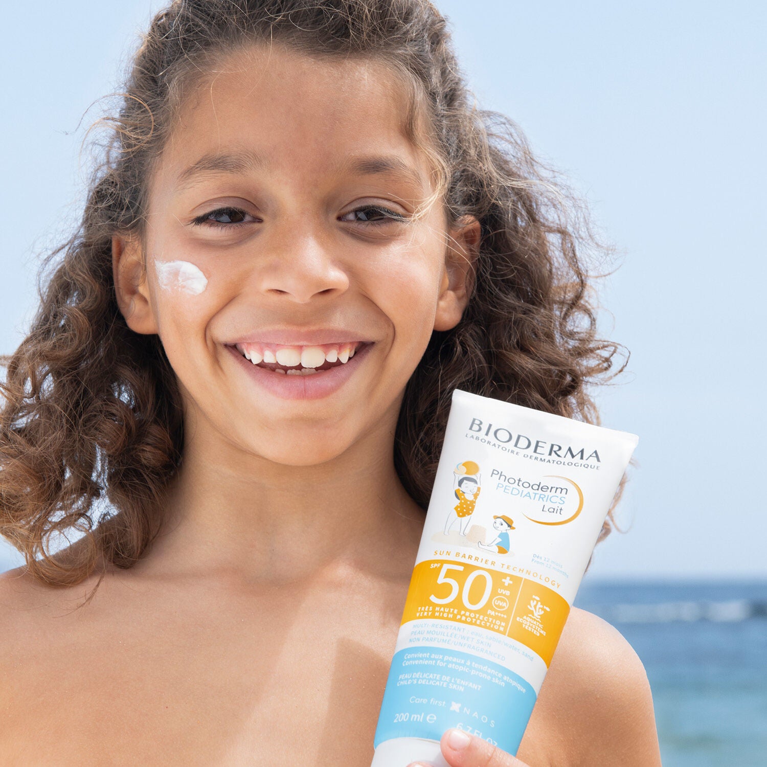 Child holding a Bioderma sunscreen tube with a beach background