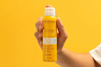 Hand holding a Bioderma sun spray bottle against a yellow background