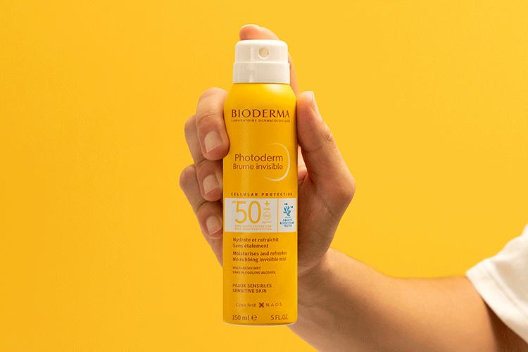 Hand holding a Bioderma sun spray bottle against a yellow background