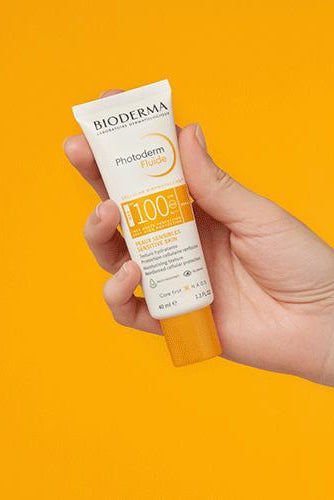 Hand holding a Bioderma sunscreen tube against an orange background